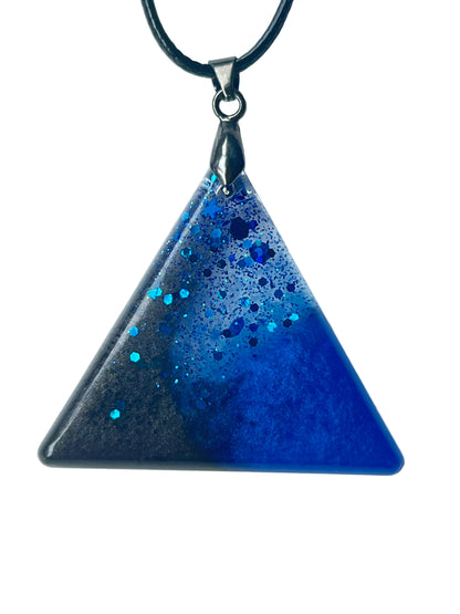 Handcrafted resin equilateral triangle pendant with deep opaque black left field and rich saturated royal-cobalt blue right field meeting diagonally, scattered with bold hexagonal glitter in electric teal and sapphire-blue throughout, suspended from a black braided cord with a gunmetal bail.