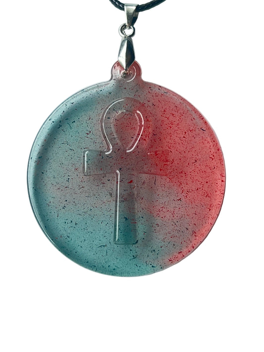 Handcrafted large resin circle pendant with soft translucent teal left field and warm coral-rose right field meeting at center, dense ultra-fine glitter throughout, and a clear raised ankh symbol embedded at center, suspended from a black braided cord with a silver-toned bail.