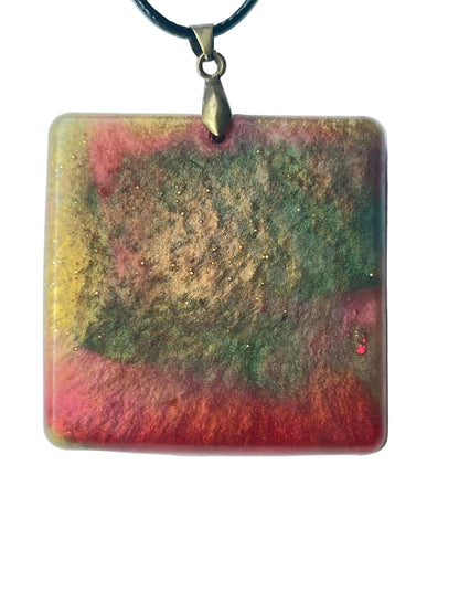 Handcrafted large resin square pendant with rich olive and forest green upper field, vivid coral-red lower bands, and a swirling golden-ochre center with fine gold glitter, suspended from a black braided cord with an antiqued brass bail.