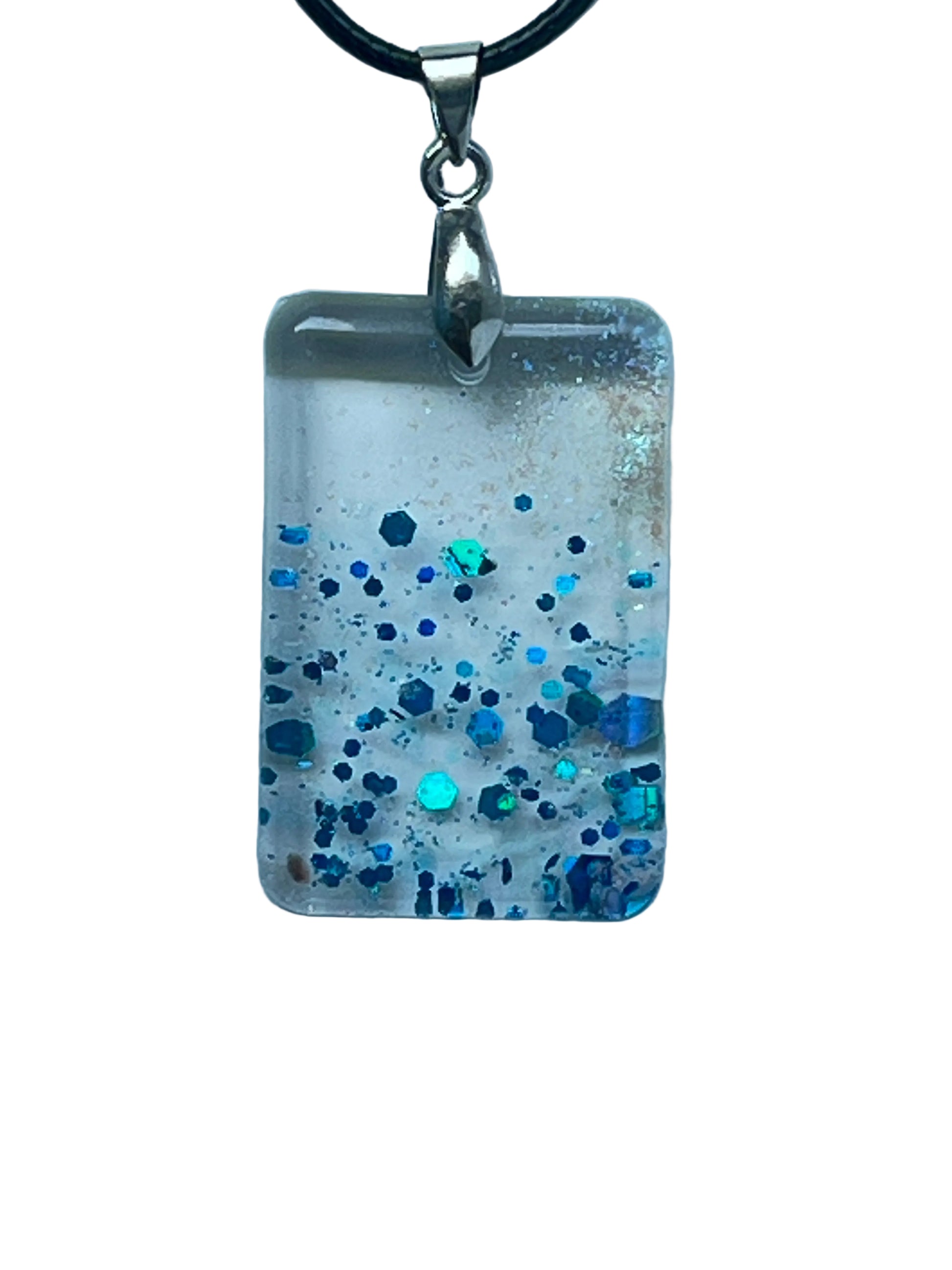 Handcrafted clear translucent resin rounded rectangle pendant with soft white wisps and bold scattered hexagonal glitter in deep sapphire navy and electric teal, suspended from a black braided cord with a silver-toned bail.