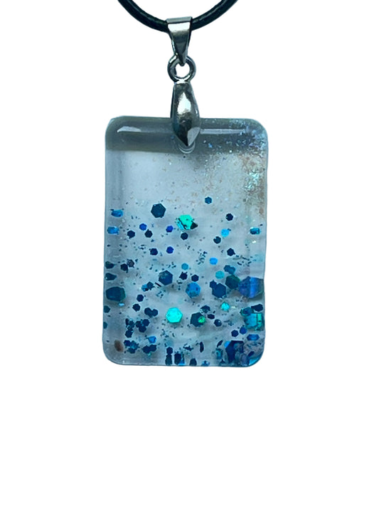 Handcrafted clear translucent resin rounded rectangle pendant with soft white wisps and bold scattered hexagonal glitter in deep sapphire navy and electric teal, suspended from a black braided cord with a silver-toned bail.