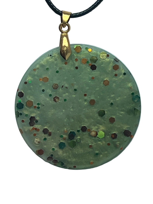Handcrafted soft pearlescent sage green resin large circle pendant with bold scattered hexagonal glitter pieces in deep burgundy, burnt orange, copper, forest green, and iridescent gold, suspended from a black braided cord with a gold-toned bail.
