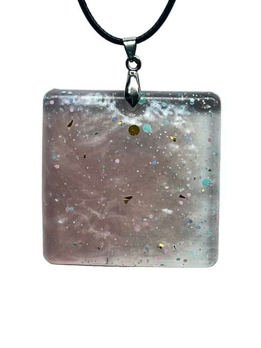 A square resin pendant with a soft dusty mauve base, milky white cloud-like swirls, and scattered confetti flecks in pink, turquoise, blue, and gold. It hangs from a silver-toned bail on a black braided cord against a pale background.