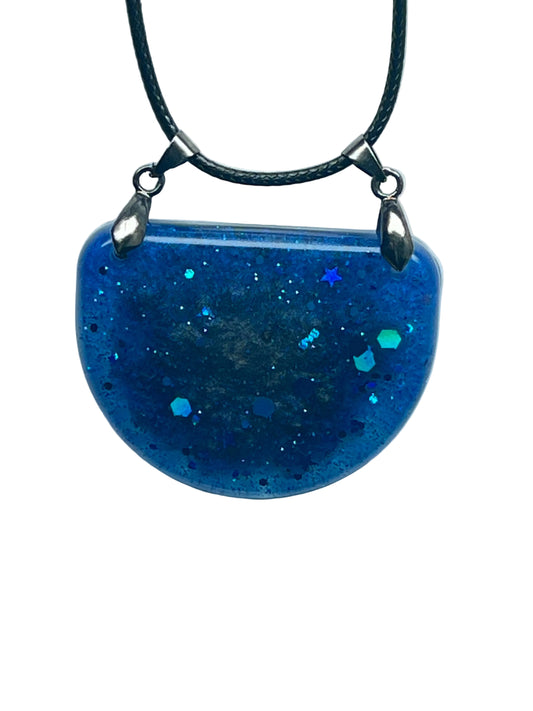 A half-circle (half-moon) shaped resin pendant in deep sapphire blue, swirled with cobalt and indigo and scattered with sparkling glitter, iridescent aqua and green hexagon flecks, and one small blue star near the top. It hangs from two gunmetal bails on a black braided cord against a pale background.