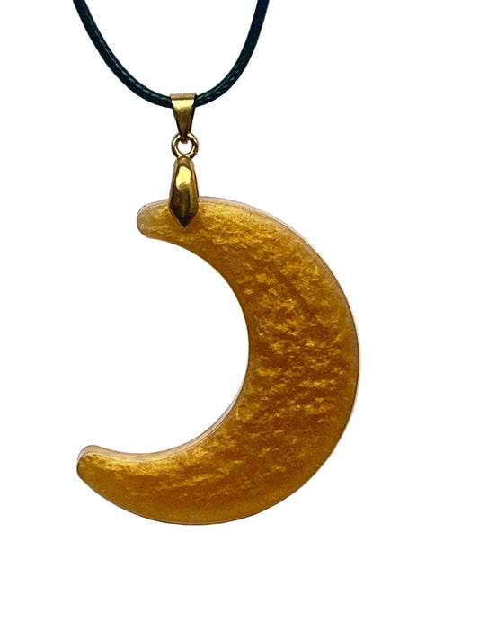 A crescent moon-shaped resin pendant in rich amber gold with a soft shimmering, beaten-metal texture. It hangs from a gold-toned bail on a black braided cord against a pale background.
