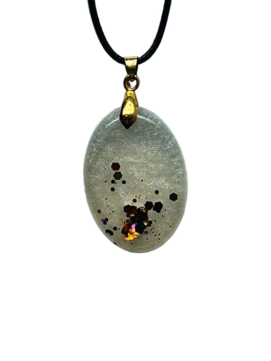 An oval resin pendant with a frosted pearl-white base and a cluster of copper, bronze, and amber hexagon flecks gathered toward the lower center, accented with a flash of iridescent gold. It hangs from a gold-toned bail on a black braided cord against a pale background.