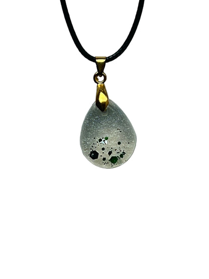 A small teardrop-shaped resin pendant with a frosted pearl-silver base and a cluster of emerald and forest-green hexagon flecks gathered toward the lower tip. It hangs from a gold-toned bail on a black braided cord against a pale background.