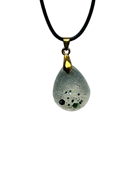 A small teardrop-shaped resin pendant with a frosted pearl-silver base and a cluster of emerald and forest-green hexagon flecks gathered toward the lower tip. It hangs from a gold-toned bail on a black braided cord against a pale background.