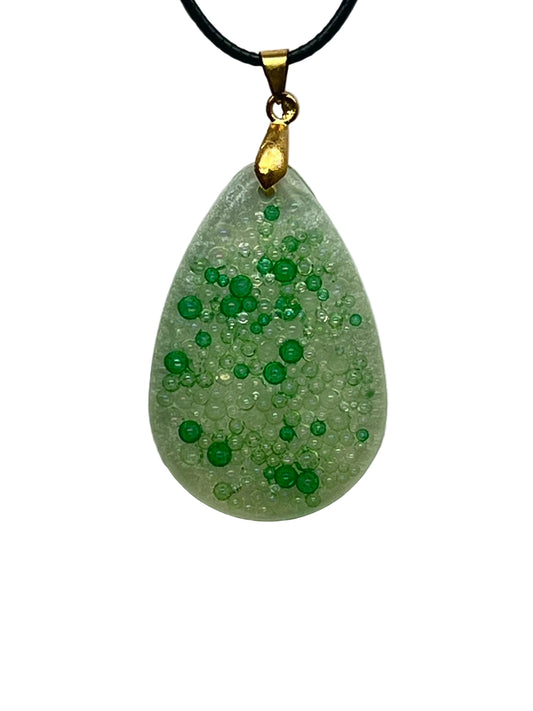 A teardrop-shaped resin pendant in seafoam green packed with tiny clear and green bubble-like beads, with clusters of brighter emerald and grass-green orbs giving a textured, fizzy appearance. It hangs from a gold-toned bail on a black braided cord against a pale background.