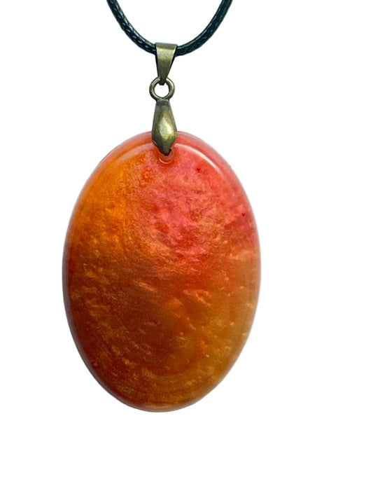 An oval-shaped resin pendant in warm swirling tones of tangerine, burnt orange, coral, and amber with a soft cloudy texture, glowing like a sunset. It hangs from an antiqued brass bail on a black braided cord against a pale background.