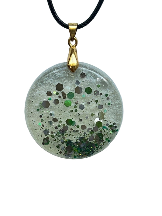 A round disc-shaped clear resin pendant scattered with confetti hexagon flecks in silver, pewter, and emerald green, denser toward the lower half with a pool of green and gold glitter near the base. It hangs from a gold-toned bail on a black braided cord against a pale background.