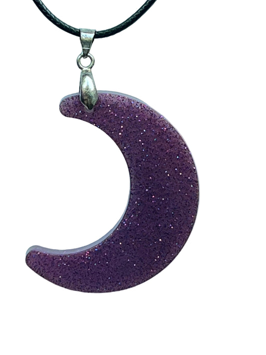 A crescent moon-shaped resin pendant in rich amethyst purple covered all over with fine sparkling glitter. It hangs from a silver-toned bail on a black braided cord against a pale background.