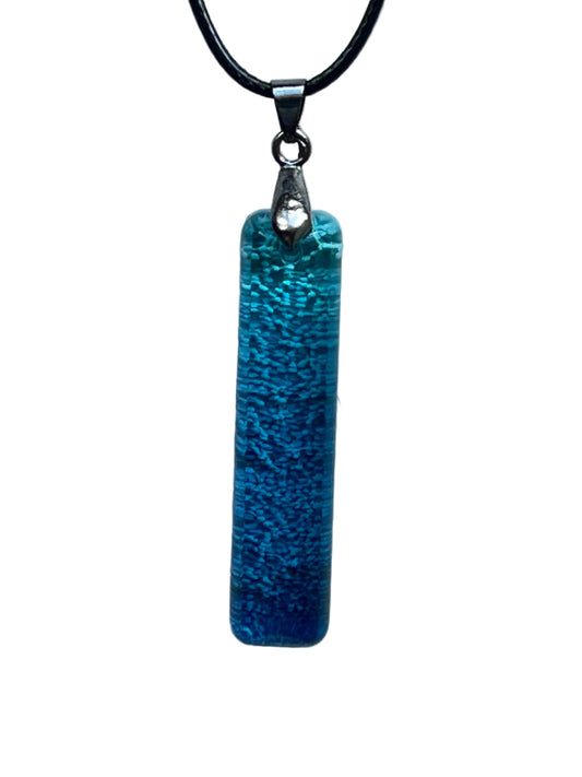 A slender vertical bar-shaped resin pendant with a finely textured, frosted surface in a gradient from bright turquoise and aqua at the top to deep ocean blue at the base. It hangs from a gunmetal bail on a black braided cord against a pale background.