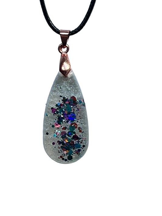 An elongated teardrop-shaped frosted clear resin pendant with a swirling ribbon of jewel-toned confetti — teal, sapphire, violet, magenta, copper, and rose-gold glitter — through the center. It hangs from a rose gold bail on a black braided cord against a pale background.