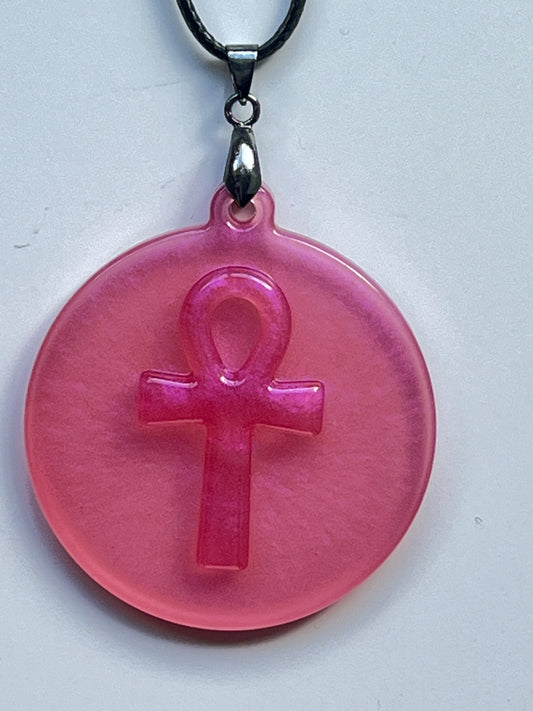A round medallion-shaped resin pendant in glowing rose pink with pearlescent shimmer and a raised, embossed ankh (Egyptian key of life) at its center. It hangs from a gunmetal bail on a black braided cord against a pale background.