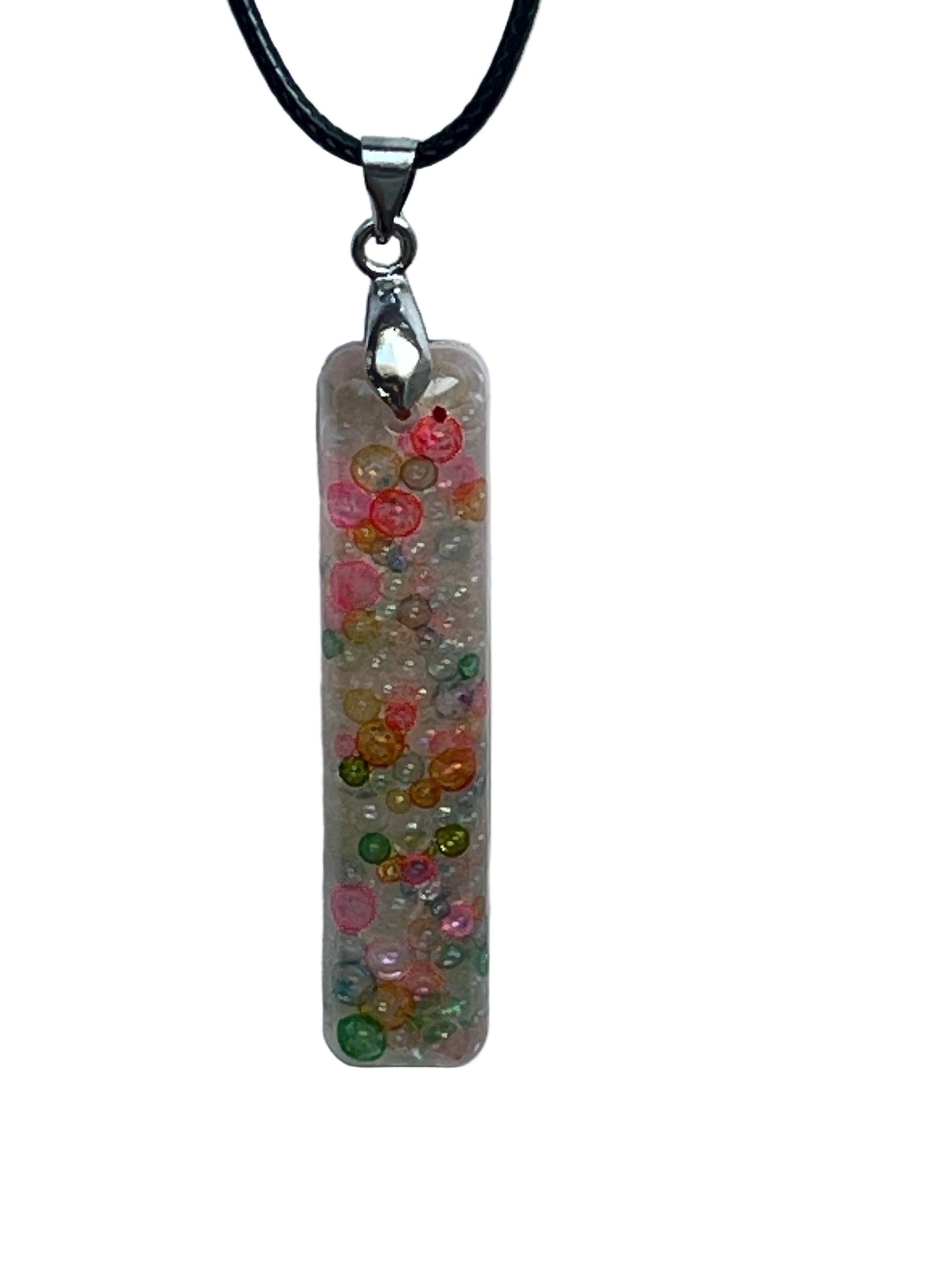 A slender vertical bar-shaped resin pendant packed with tiny bubble-beads and micro-pearls in pastel confetti colors — coral, peach, gold, mint green, and pink — with a soft pearly shimmer. It hangs from a silver-toned bail on a black braided cord against a pale background.
