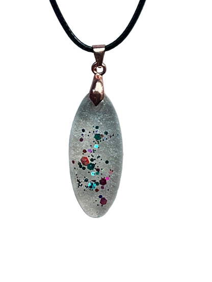 An elongated marquise-shaped frosted clear resin pendant with a ribbon of multicolored confetti flecks — green, red, orange, pink, and aqua with gold sparkle — scattered down the center. It hangs from a rose gold bail on a black braided cord against a pale background.