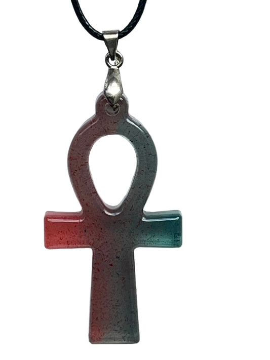 An ankh-shaped (Egyptian cross with a looped top) resin pendant with a gradient from warm coral and red on the left to cool teal and green on the right, scattered with fine dark speckles. It hangs from a silver-toned bail on a black braided cord against a pale background.