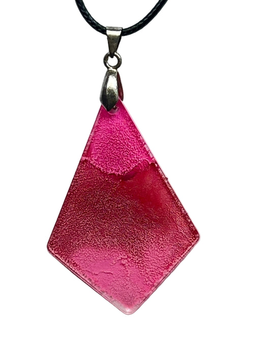 A kite-shaped (elongated diamond) resin pendant in warm rose pink with a soft suede-like texture, deepening to coral and rosy-gold in the center. It hangs from a silver-toned bail on a black braided cord against a pale background.