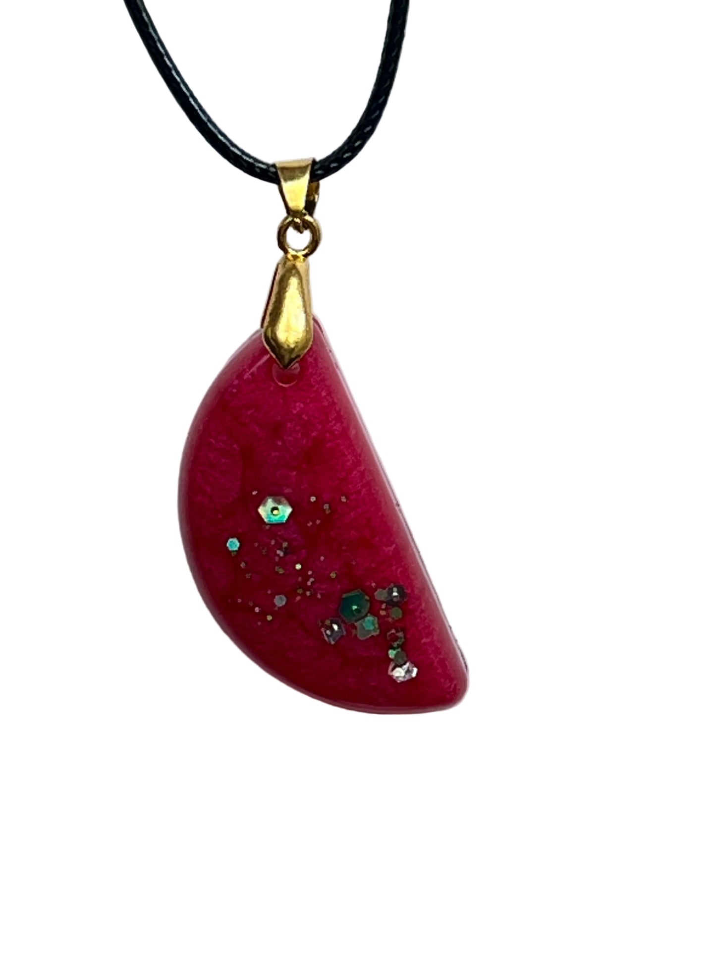 A half-circle (half-moon) shaped resin pendant in warm rose red with pearlescent shimmer and a low cluster of iridescent green, silver, and gold confetti hexagon flecks. It hangs from a gold-toned bail on a black braided cord against a pale background.