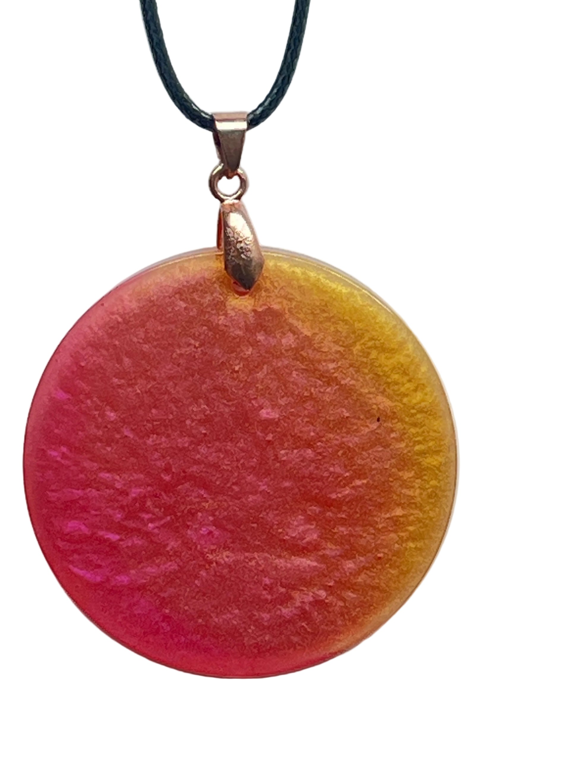 A round disc-shaped resin pendant with a pearlescent gradient sweeping from pink and magenta on one side to golden-yellow on the other, with a frosted, luminous texture. It hangs from a rose gold bail on a black braided cord against a pale background.