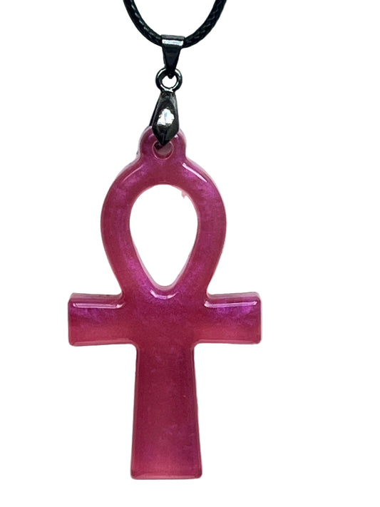 An ankh-shaped (Egyptian cross with a looped top) resin pendant in a glowing rose pink with pearlescent shimmer and deeper magenta tones. It hangs from a gunmetal bail on a black braided cord against a pale background.