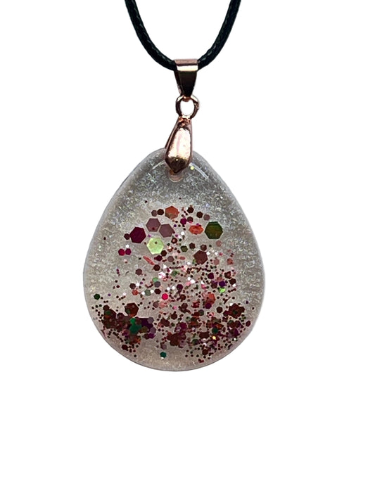 A teardrop-shaped frosted clear resin pendant with a burst of multicolored confetti — copper, rose-gold, ruby, lime green, fuchsia, and silver glitter — clustered through the lower half. It hangs from a rose gold bail on a black braided cord against a pale background.