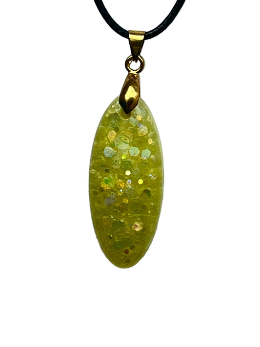 An elongated oval-shaped resin pendant in soft buttery yellow scattered throughout with iridescent gold, green, and silver confetti hexagon flecks. It hangs from a gold-toned bail on a black braided cord against a pale background.