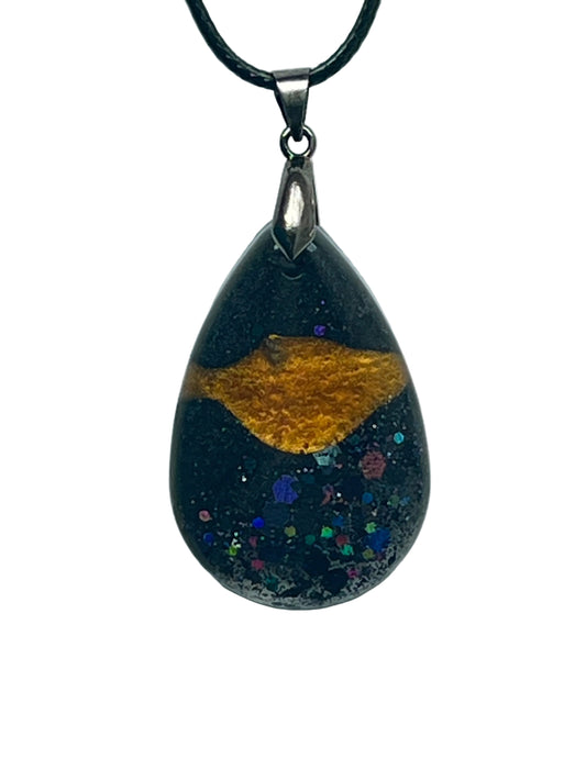 A teardrop-shaped resin pendant with a deep black base, a streak of molten gold across the upper half, and a scatter of iridescent rainbow confetti hexagons across the lower half like stars. It hangs from a gunmetal bail on a black braided cord against a pale background.
