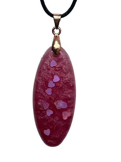 An elongated oval-shaped resin pendant in dusty mauve-rose with a frosted texture and a trail of small lilac and iridescent heart-shaped confetti drifting down the center. It hangs from a rose gold bail on a black braided cord against a pale background.