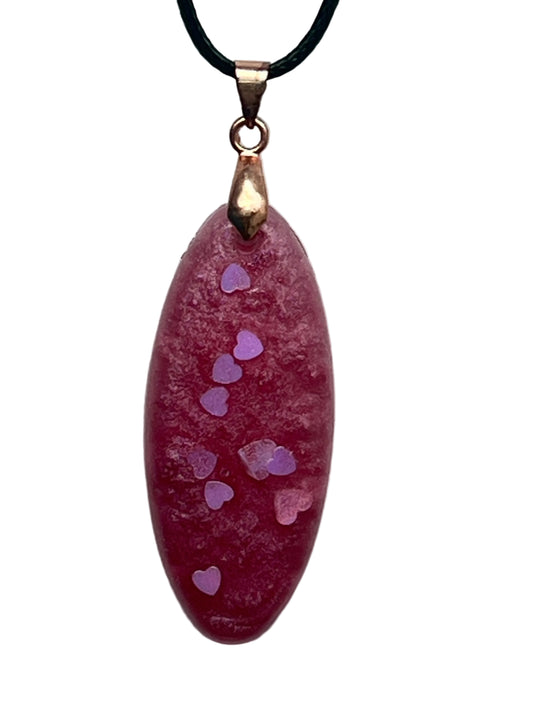 An elongated oval-shaped resin pendant in dusty mauve-rose with a frosted texture and a trail of small lilac and iridescent heart-shaped confetti drifting down the center. It hangs from a rose gold bail on a black braided cord against a pale background.