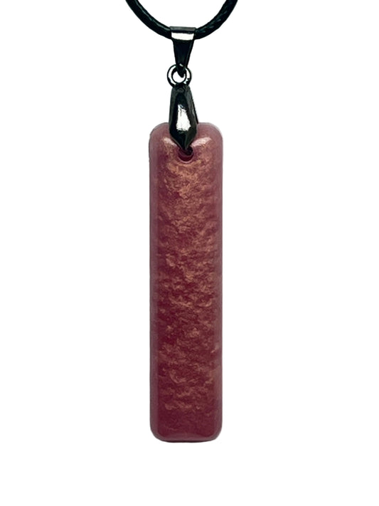 A slender vertical bar-shaped resin pendant in warm mauve-rose with a velvety suede-like texture and coppery, peach-toned mottling throughout. It hangs from a gunmetal bail on a black braided cord against a pale background.
