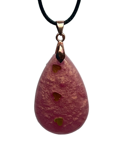 A teardrop-shaped resin pendant in dusty rose pink with a frosted texture and warm coppery-gold tones, with three gold heart-shaped confetti pieces lined up vertically down the center. It hangs from a rose gold bail on a black braided cord against a pale background.