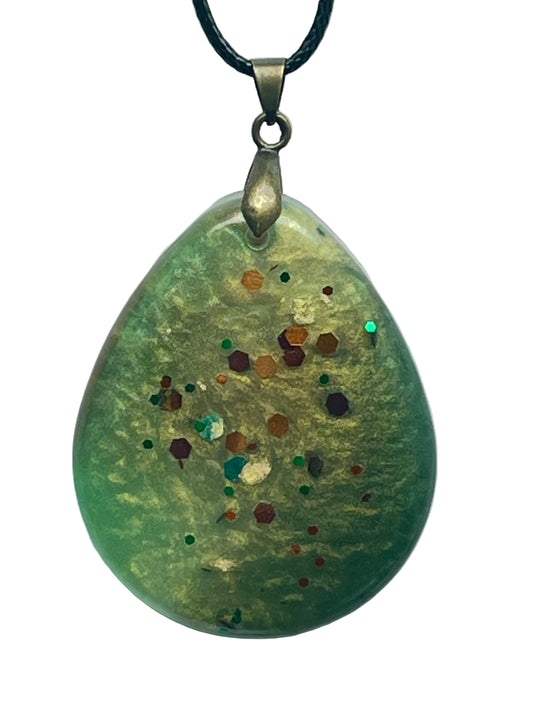 A teardrop-shaped resin pendant in pearlescent sage-green and gold, scattered with confetti hexagon flecks in copper, rust, burgundy, teal, and emerald. It hangs from an antiqued brass bail on a black braided cord against a pale background.