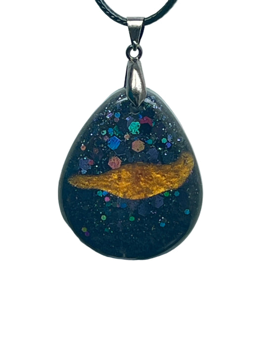 A teardrop-shaped resin pendant with a deep glittering black base, a streak of molten gold across the center like a comet, and scattered iridescent violet, blue, teal, pink, and green confetti hexagons like nebulae. It hangs from a gunmetal bail on a black braided cord against a pale background.
