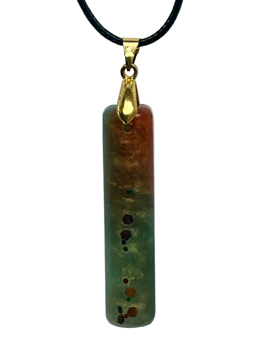 A slender vertical bar-shaped resin pendant with a gradient from warm rust and orange at the top through golden amber to sage green at the base, with a vertical trail of burgundy, copper, and emerald confetti flecks down the center. It hangs from a gold-toned bail on a black braided cord against a pale background.