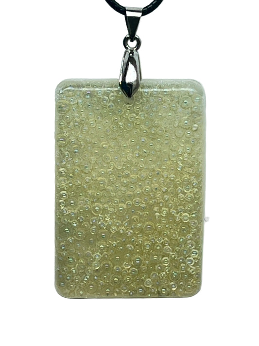 A rectangular resin pendant in creamy ivory and pale gold, packed completely full of tiny iridescent bubble-beads and micro-pearls with a soft opal shimmer. It hangs from a silver-toned bail on a black braided cord against a pale background.