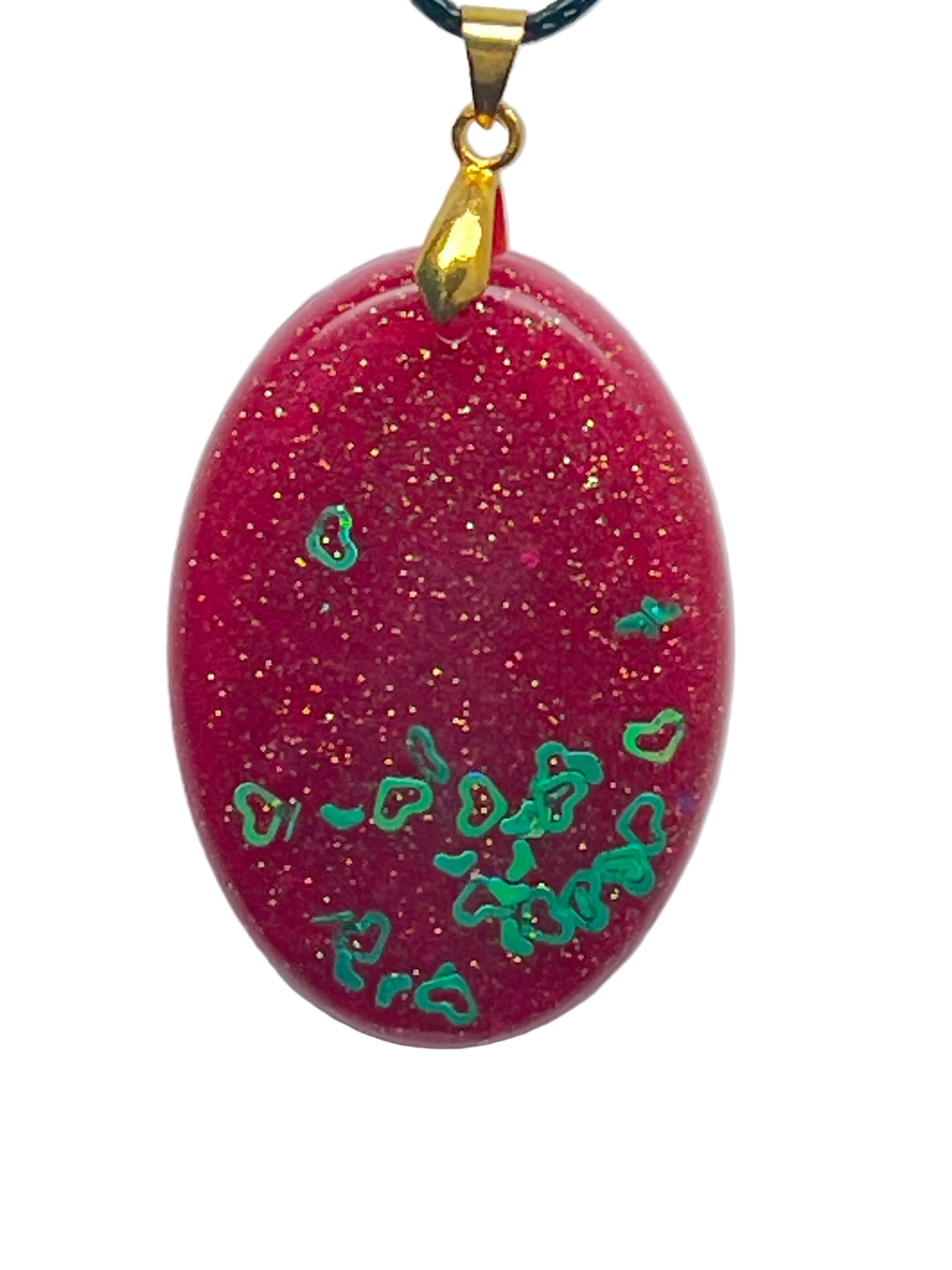 An oval-shaped resin pendant in glittering ruby red with fine gold and orange sparkle, scattered across the lower half with iridescent green heart-shaped confetti. It hangs from a gold-toned bail on a black braided cord against a pale background.