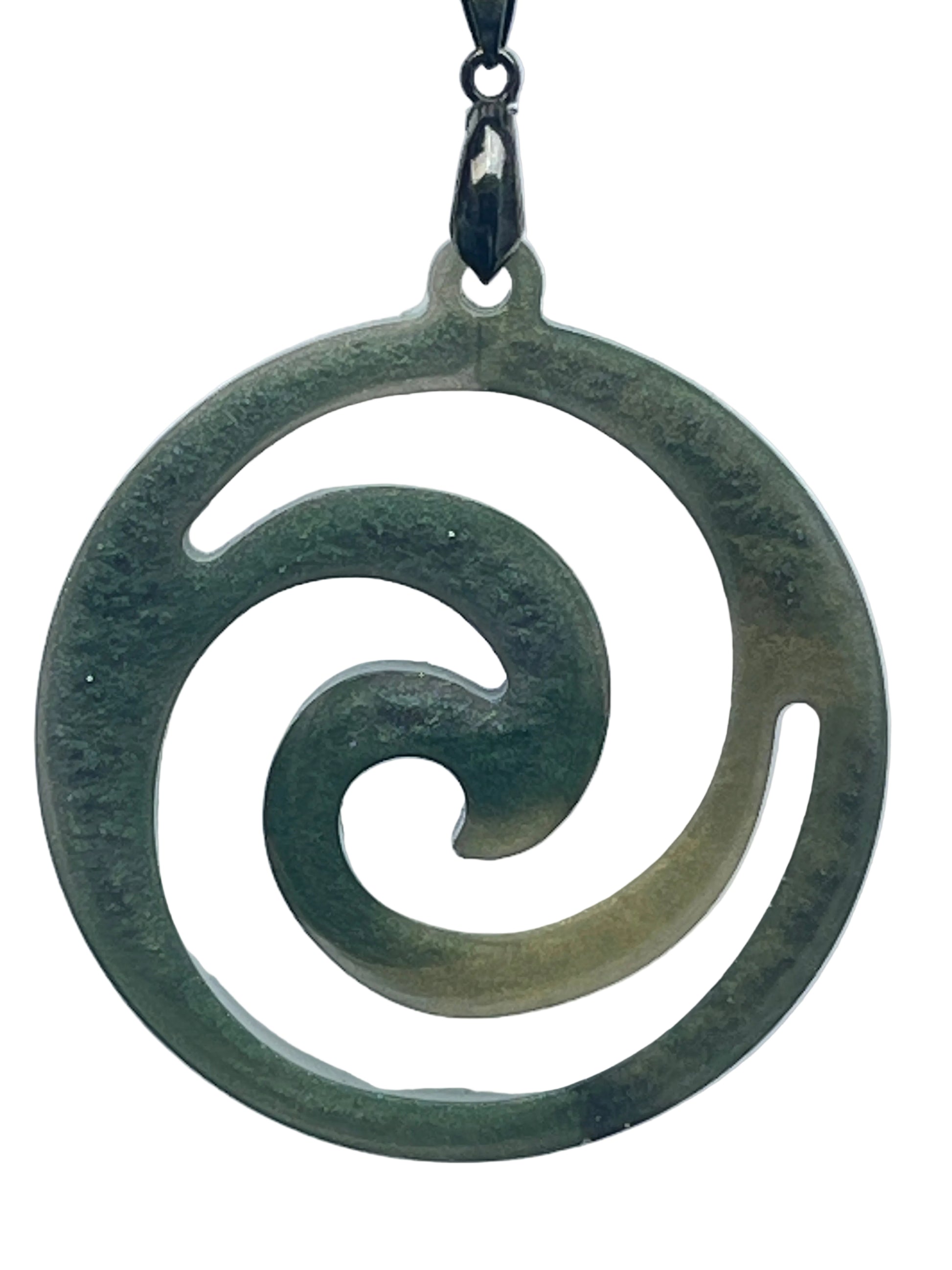 A round resin pendant in mottled forest and sage green resembling carved jade, featuring an open koru spiral cut-out design that curls inward from the outer ring toward the center. It hangs from a gunmetal bail on a black braided cord against a pale background.