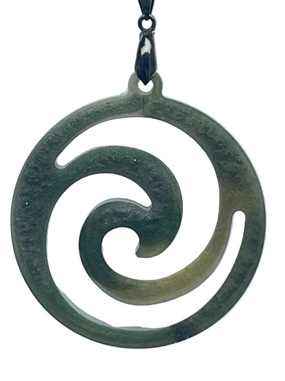 A round resin pendant in mottled forest and sage green resembling carved jade, featuring an open koru spiral cut-out design that curls inward from the outer ring toward the center. It hangs from a gunmetal bail on a black braided cord against a pale background.