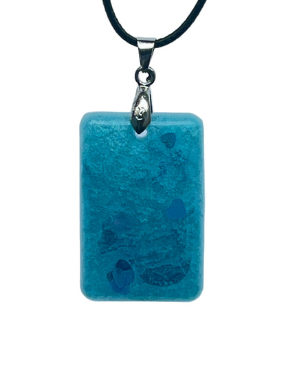 A rectangular resin pendant in frosted aqua and teal blue with a mottled, watery texture, scattered across the lower half with blue and iridescent heart-shaped confetti. It hangs from a silver-toned bail on a black braided cord against a pale background.