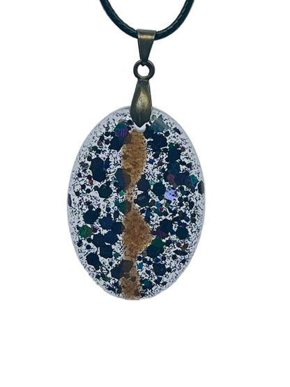 An oval-shaped resin pendant with a frosted clear, silver-glittered base densely scattered with dark black, teal, violet, and iridescent green speckles like a starfield, with a warm amber-gold vein winding down the center. It hangs from an antiqued brass bail on a black braided cord against a pale background.