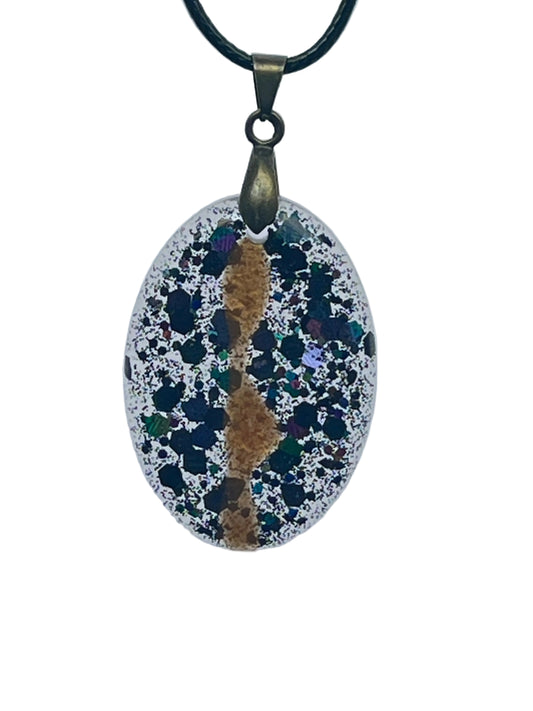 An oval-shaped resin pendant with a frosted clear, silver-glittered base densely scattered with dark black, teal, violet, and iridescent green speckles like a starfield, with a warm amber-gold vein winding down the center. It hangs from an antiqued brass bail on a black braided cord against a pale background.