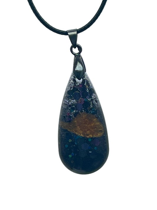A teardrop-shaped resin pendant in deep navy-black with scattered iridescent violet, teal, and silver speckle, and a single warm amber-gold shape drifting near the center. It hangs from a gunmetal bail on a black braided cord against a pale background.