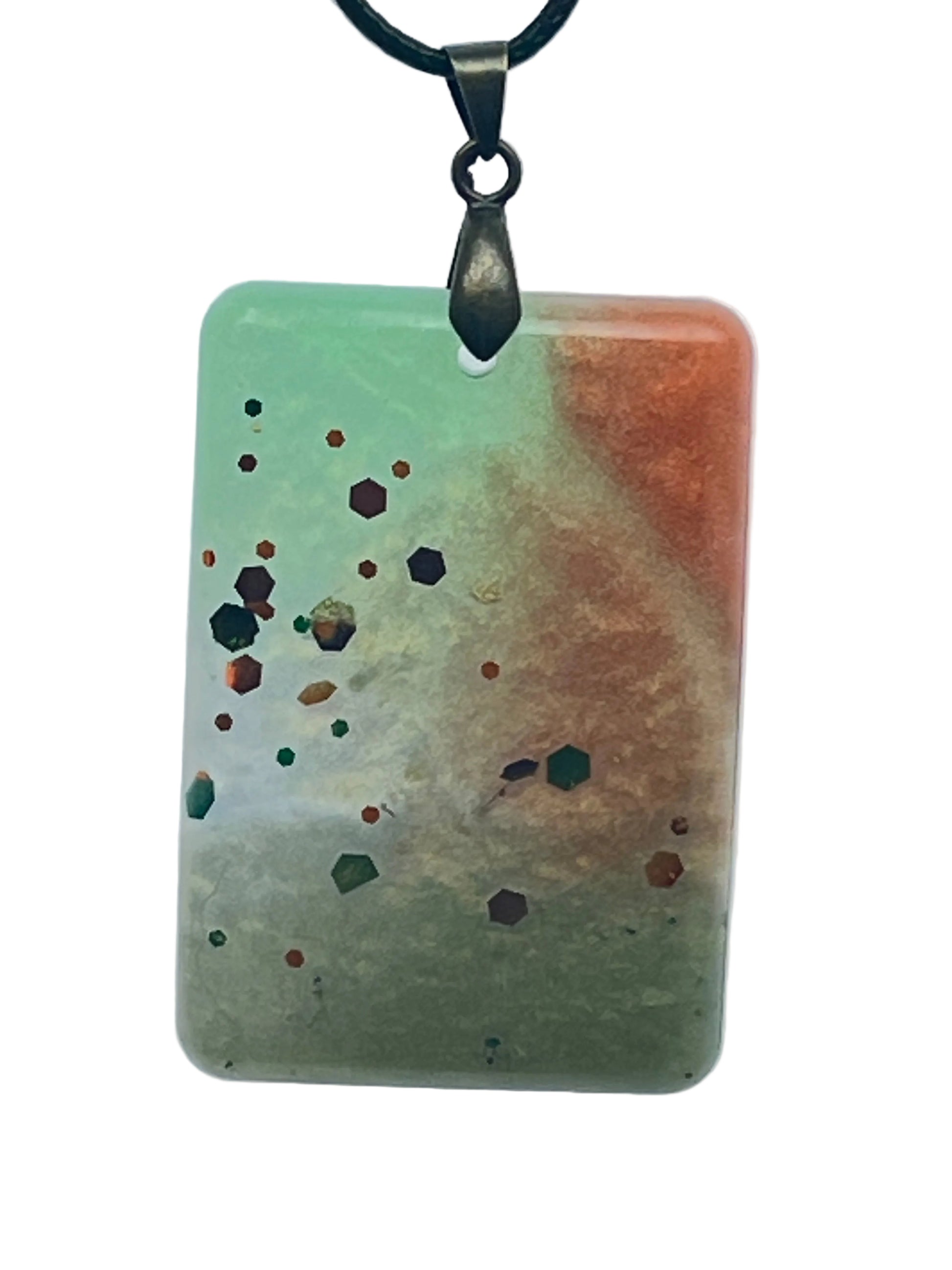 A rectangular resin pendant with a diagonal gradient from soft mint green to warm coral and rust, scattered with confetti hexagon flecks in burgundy, copper, and forest green. It hangs from an antiqued brass bail on a black braided cord against a pale background.