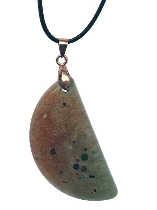 An organically curved, freeform resin pendant with a gradient from umber brown at the top to sage green at the base, scattered with confetti hexagon flecks in emerald, plum, and forest green. It hangs from a rose gold bail on a black braided cord against a pale background.