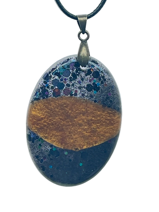 An oval-shaped resin pendant with a bold band of molten amber-gold across the center, framed above and below by dense cosmic speckle in black, violet, teal, and silver. It hangs from an antiqued brass bail on a black braided cord against a pale background.