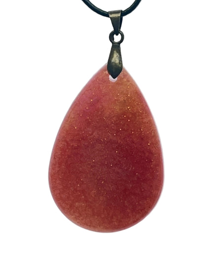 A full, rounded teardrop-shaped resin pendant in deep rust red with warm coral undertones near the top, scattered throughout with fine gold sparkle. It hangs from an antiqued brass bail on a black braided cord against a pale background.