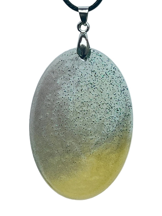 A large oval-shaped resin pendant with a gradient from soft grey with fine glitter at the top to pale golden-yellow at the base. It hangs from a silver-toned bail on a black braided cord against a pale background.