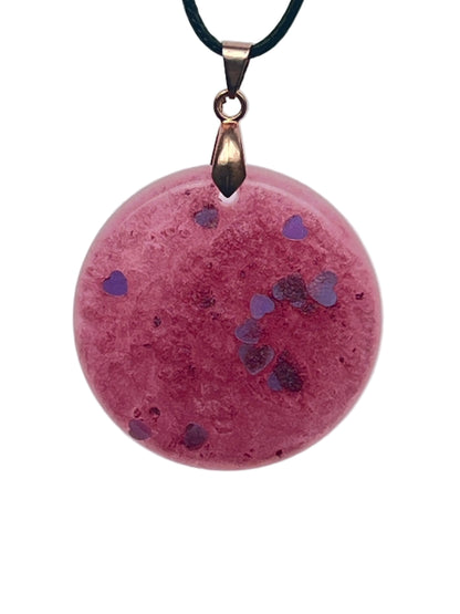 A round disc-shaped resin pendant in dusty mauve-pink with a mottled, frosted texture, scattered diagonally with tiny violet and periwinkle heart-shaped confetti. It hangs from a rose gold bail on a black braided cord against a pale background.